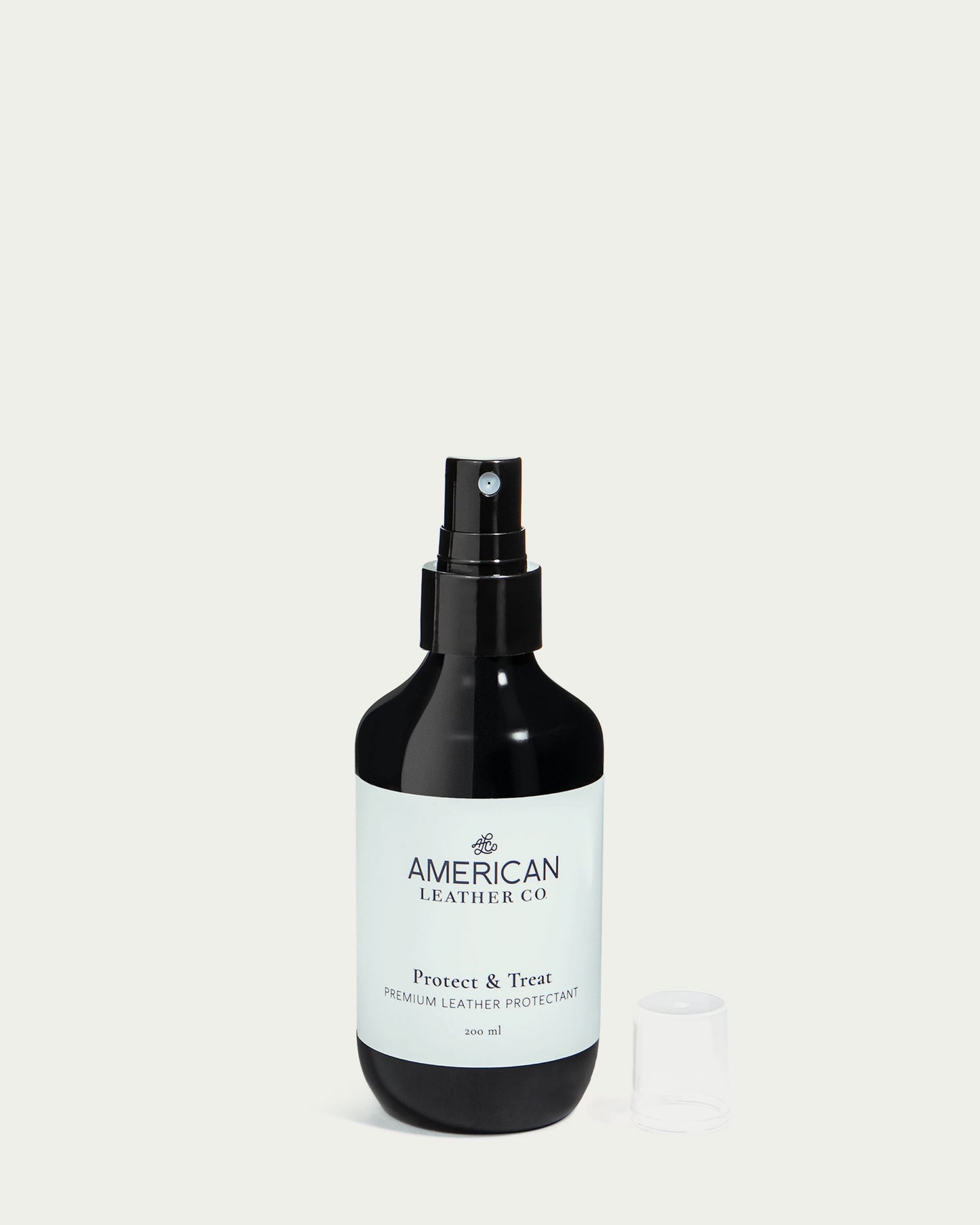 A dark green spray bottle labeled "Protect & Treat Premium Leather Protectant Spray" from American Leather Co. stands on a light background. The leather treatment effectively repels water with its stain-resistant formula, and a clear plastic cap is neatly placed beside it.