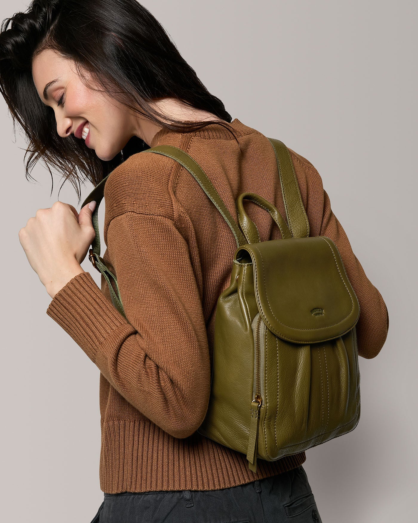 Woman smiling and looking down, wearing a brown sweater and carrying an olive green leather backpack over her shoulders against a neutral background.