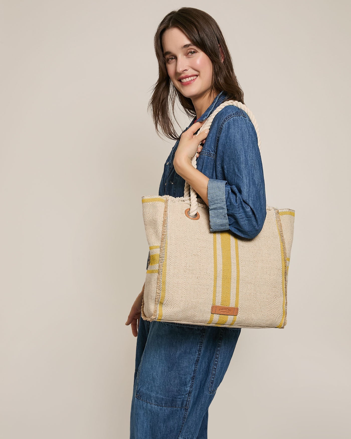 Woman in a denim jumpsuit smiles while holding a beige woven tote bag with yellow stripes and rope handles over her shoulder, standing against a plain light background.