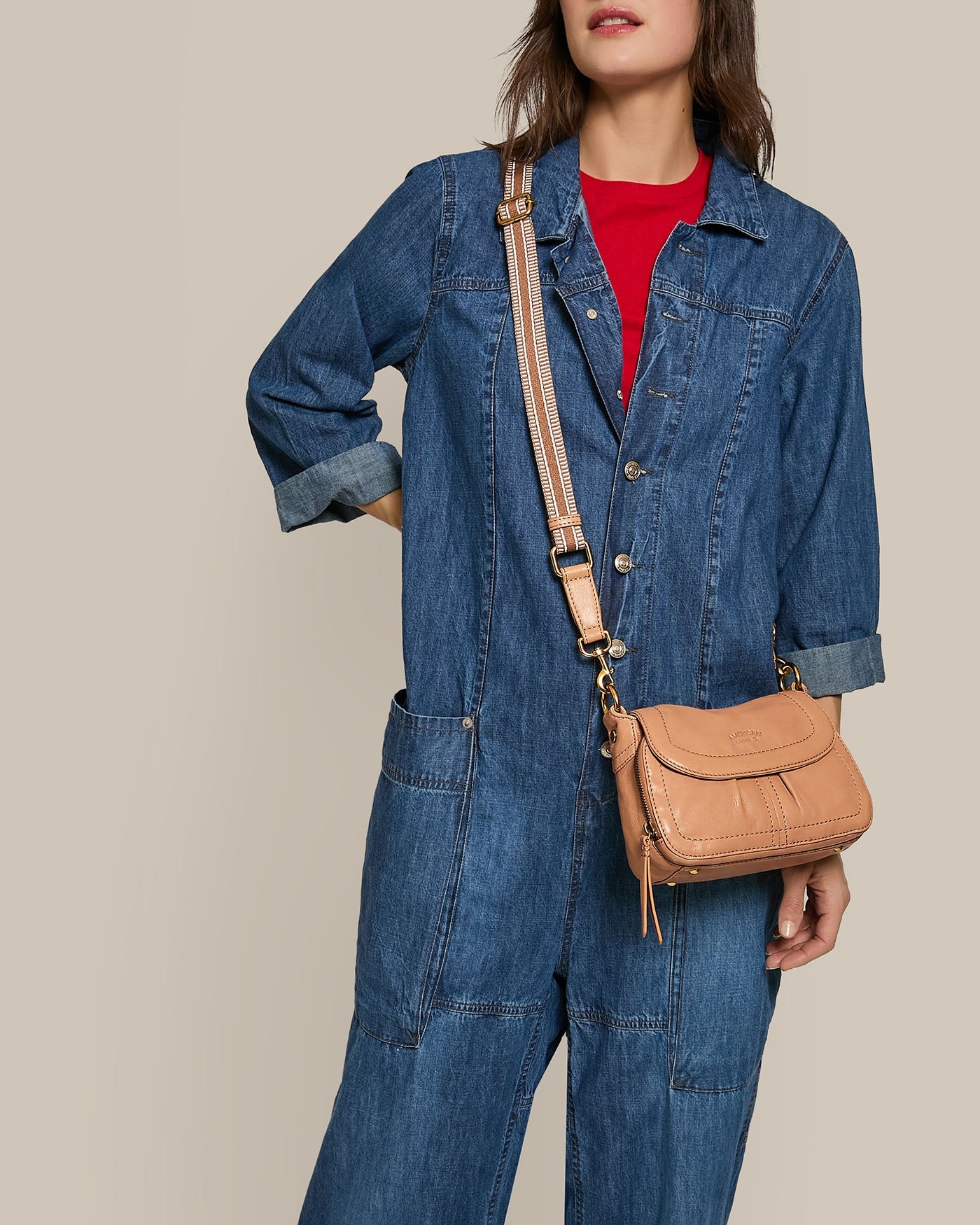 A person in a denim jumpsuit with rolled-up sleeves, wearing a red shirt underneath, and carrying a tan leather crossbody bag. The background is a plain beige.