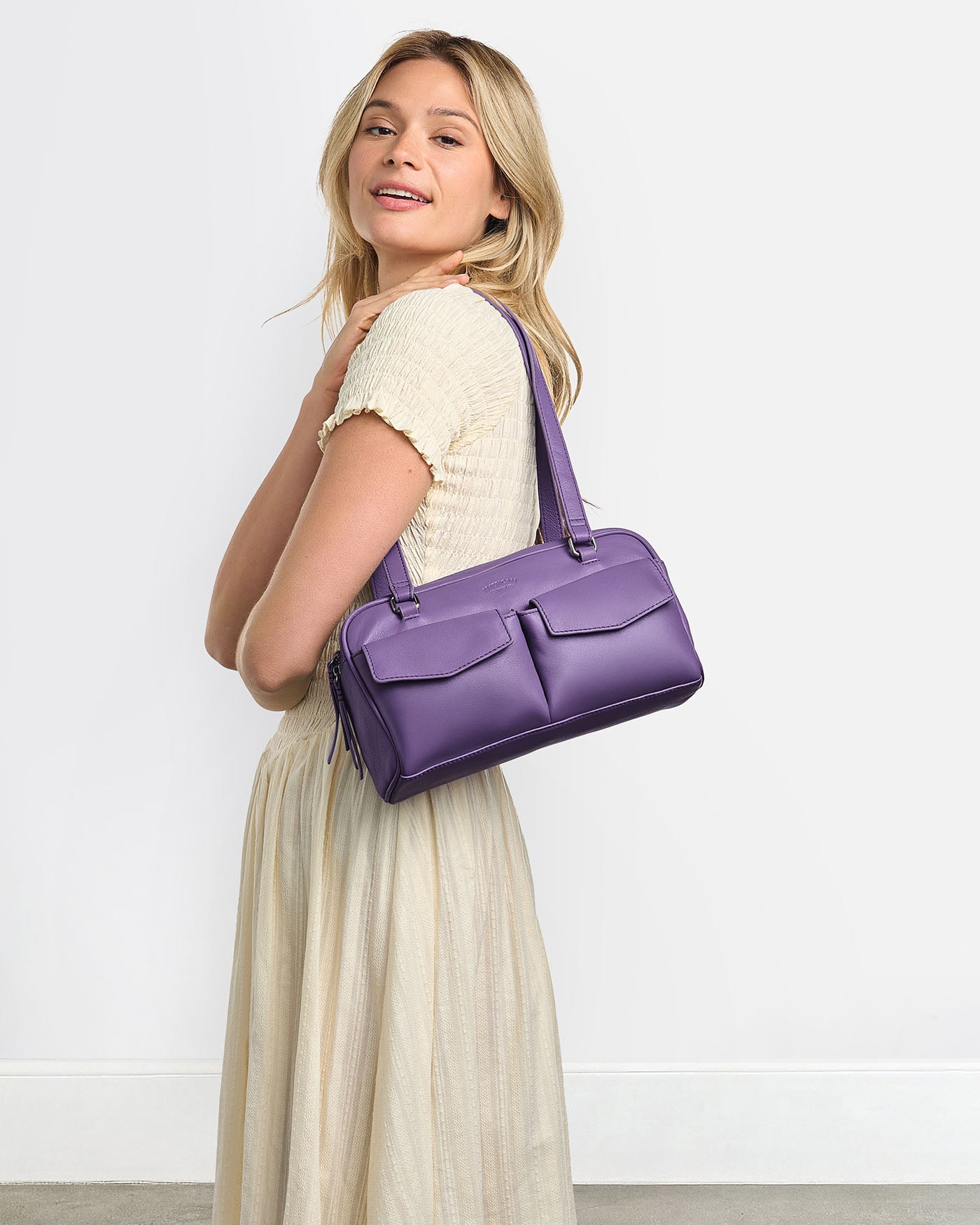 A woman in a cream dress stands smiling, holding a purple handbag with two front pockets over her shoulder against a plain light background.