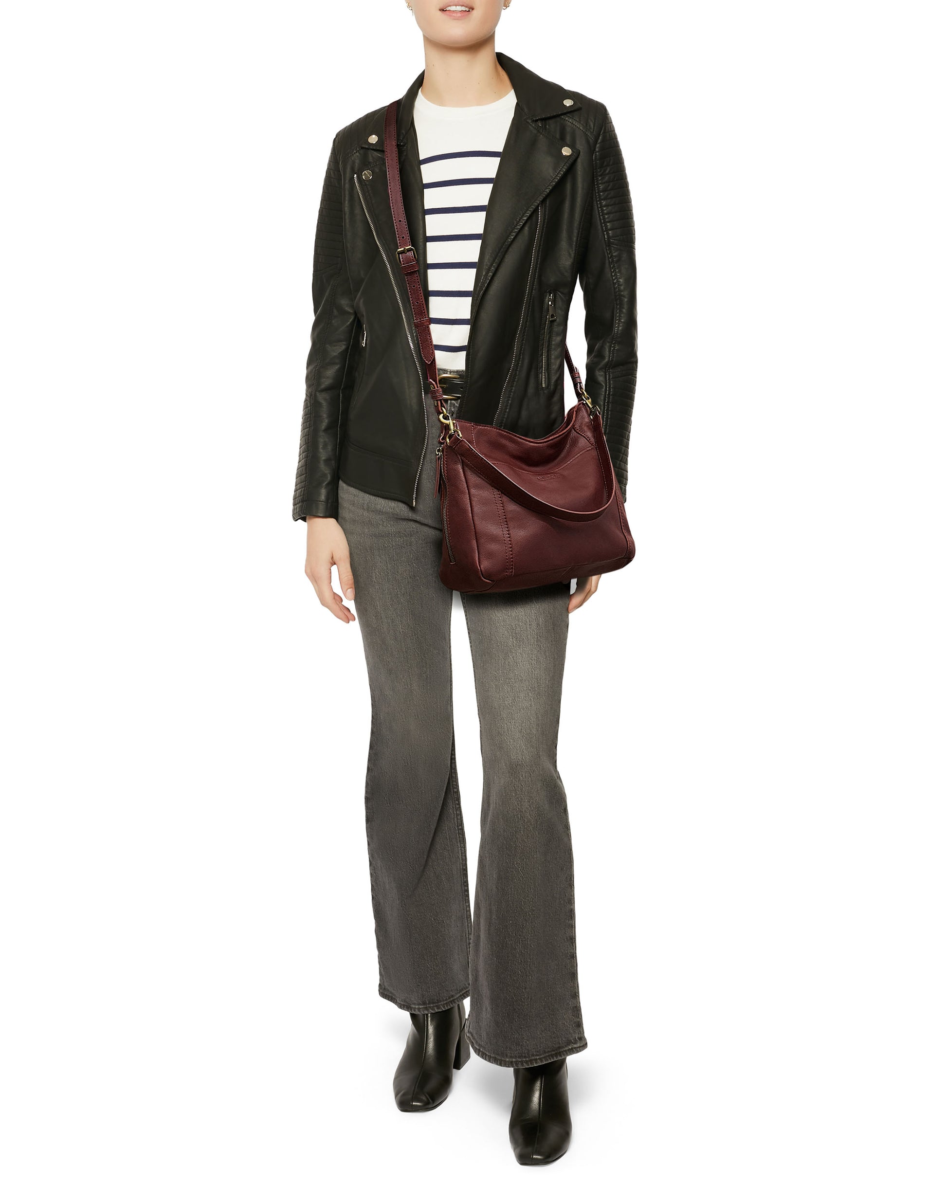 A person wearing a black leather jacket, a striped shirt, gray flared jeans, and black ankle boots carries the Austin Convertible Shoulder Crossbody by American Leather Co., featuring side zipper pockets in maroon. The outfit is casual and stylish.