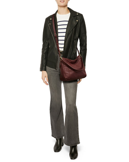 A person wearing a black leather jacket, a striped shirt, gray flared jeans, and black ankle boots carries the Austin Convertible Shoulder Crossbody by American Leather Co., featuring side zipper pockets in maroon. The outfit is casual and stylish.