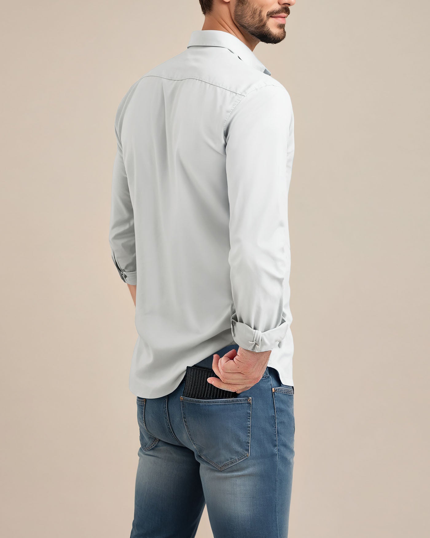 A bearded man in a light gray long-sleeve shirt and blue jeans stands with his back to the camera. His left hand in a pocket conceals the Shorewood Bifold Wallet by American Leather Co. against a simple beige background.