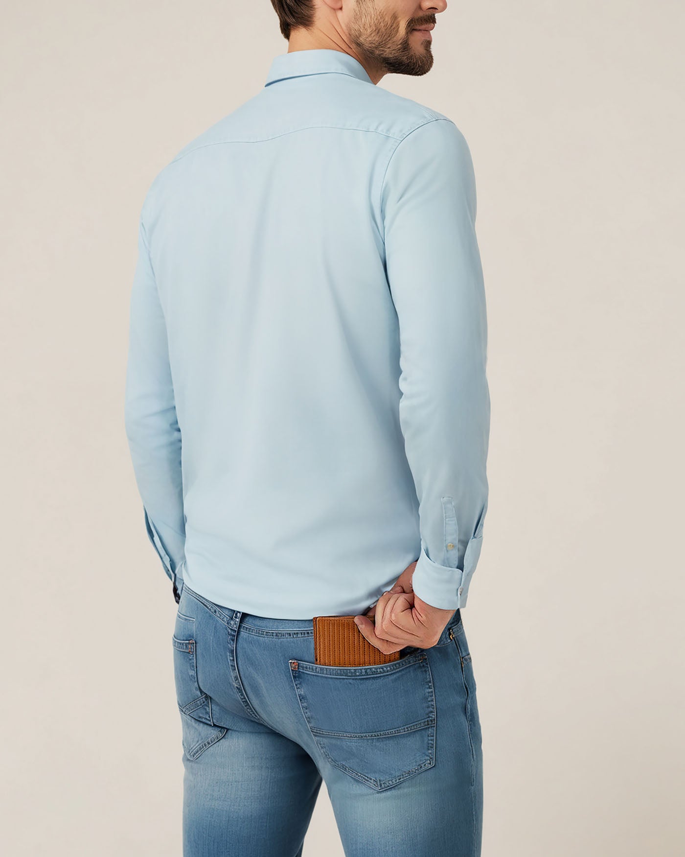A man in a light blue shirt and jeans is seen from behind, slipping an American Leather Co. Shorewood Bifold Wallet made of premium nappa leather into his back pocket.