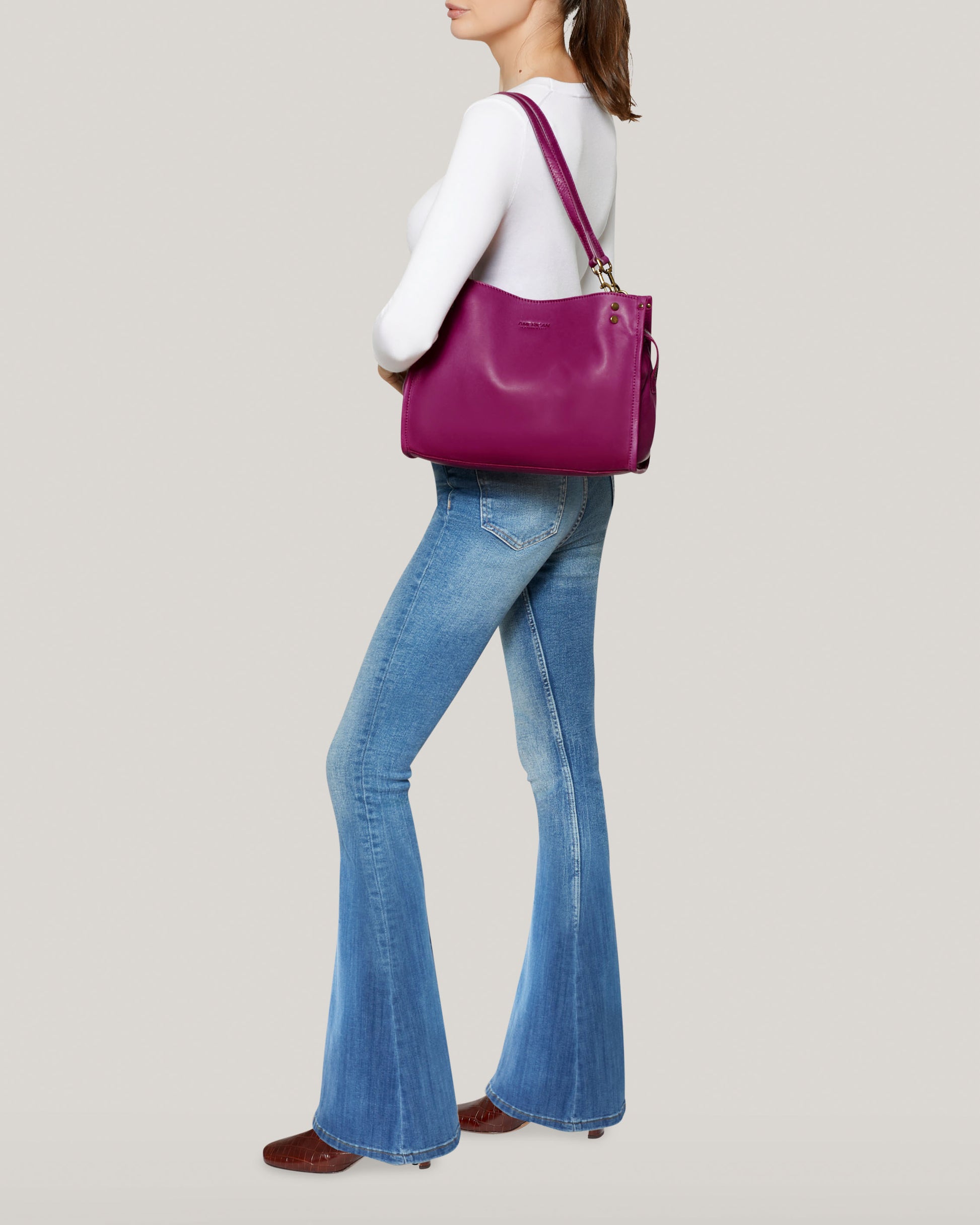 A person in a white long-sleeve top and blue flared jeans stands in profile view, holding the Lenox Triple Entry Satchel by American Leather Co. They wear dark shoes with their hair pulled back. The plain, light-colored background accentuates this everyday essential look.
