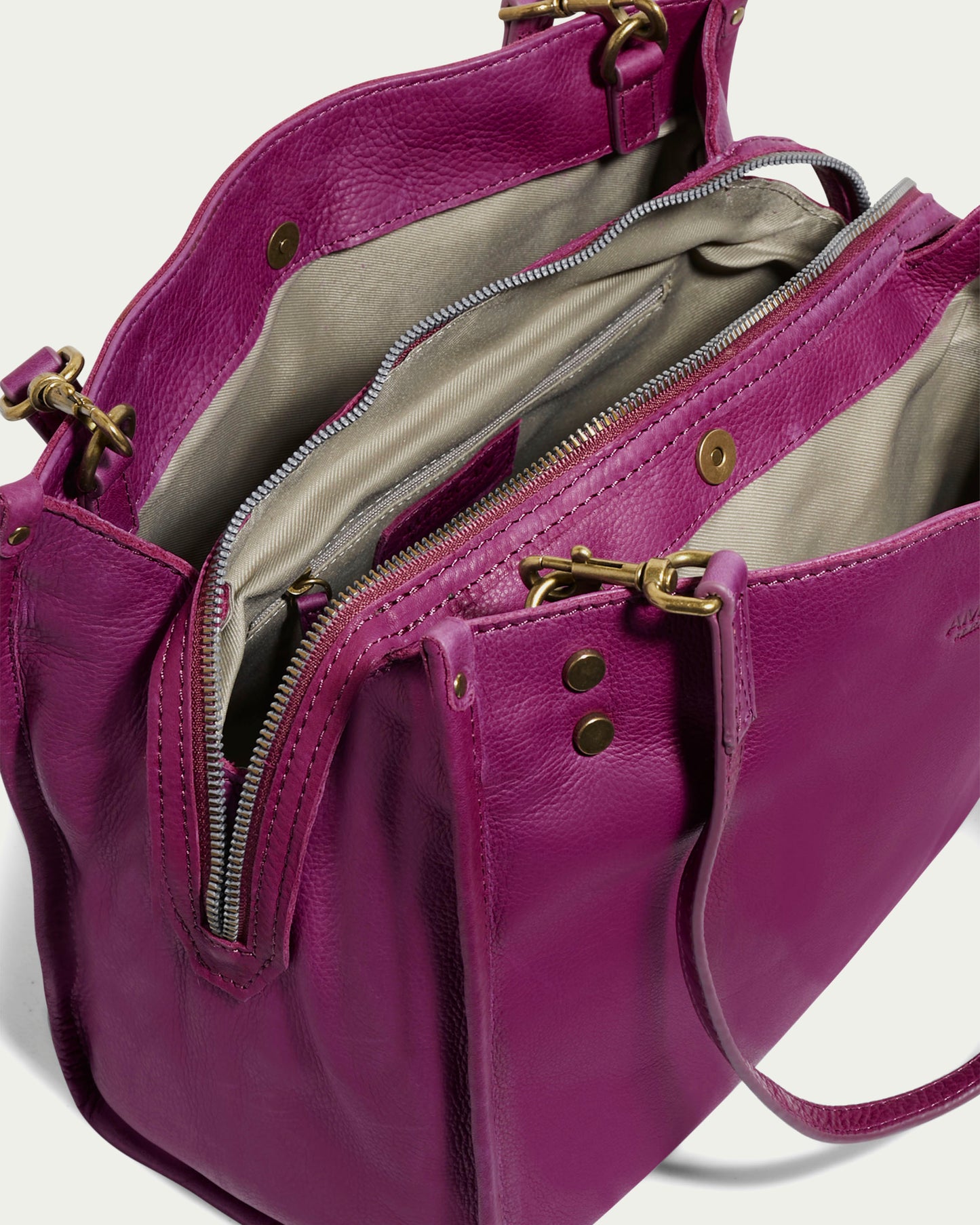 Introducing the Lenox Triple Entry Satchel from American Leather Co., this chic handbag is crafted from genuine American leather in a sophisticated purple hue. It boasts multiple compartments and opens to exhibit beige interior linings complemented by brass zippers. The satchel is designed with two handles enhanced by detailed brass hardware and rivets, making it an everyday essential.
